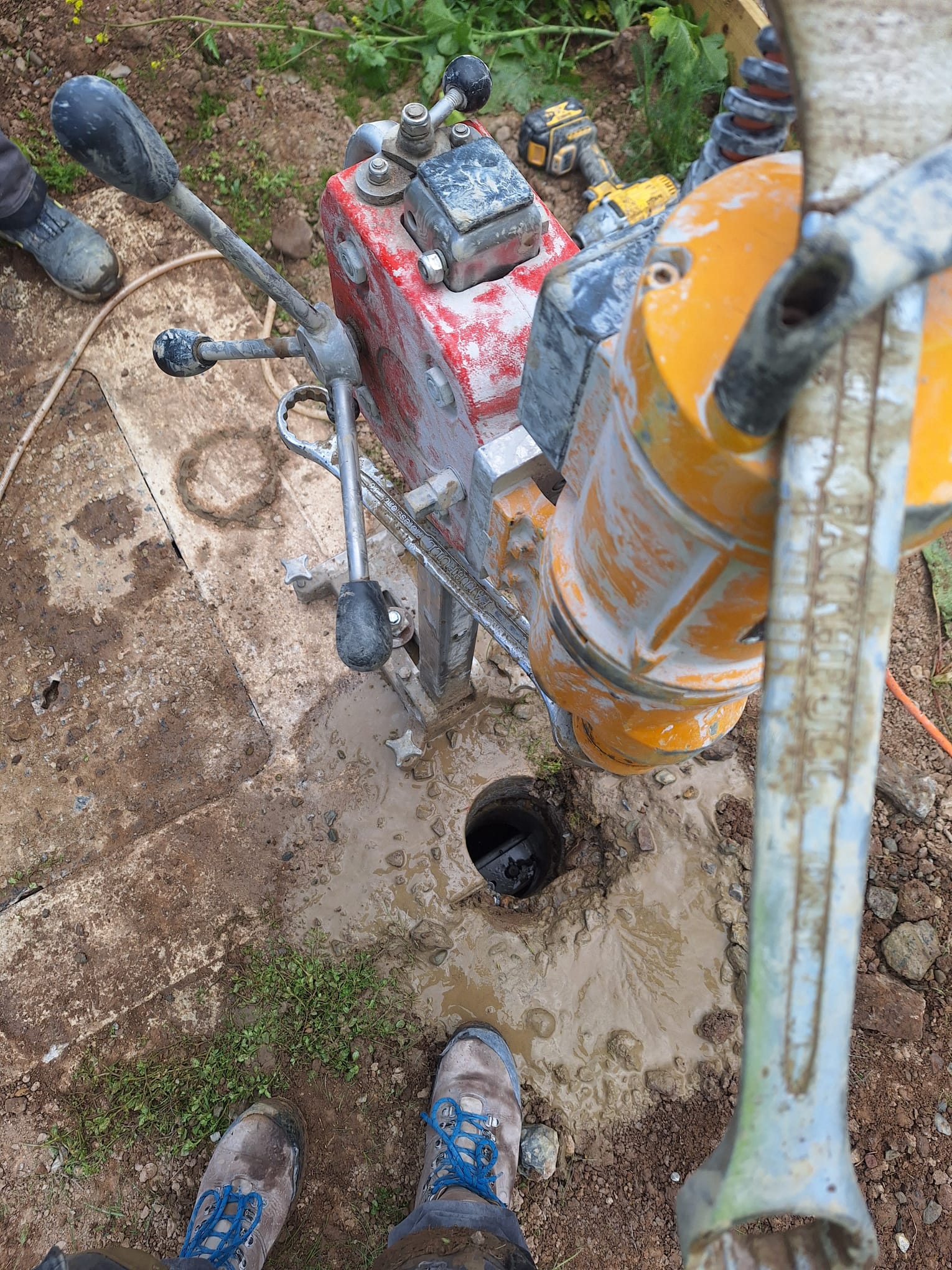 Rig-mounted diamond core drill boring through outdoor concrete slab for manhole access — operator in hi-vis, water suppression active